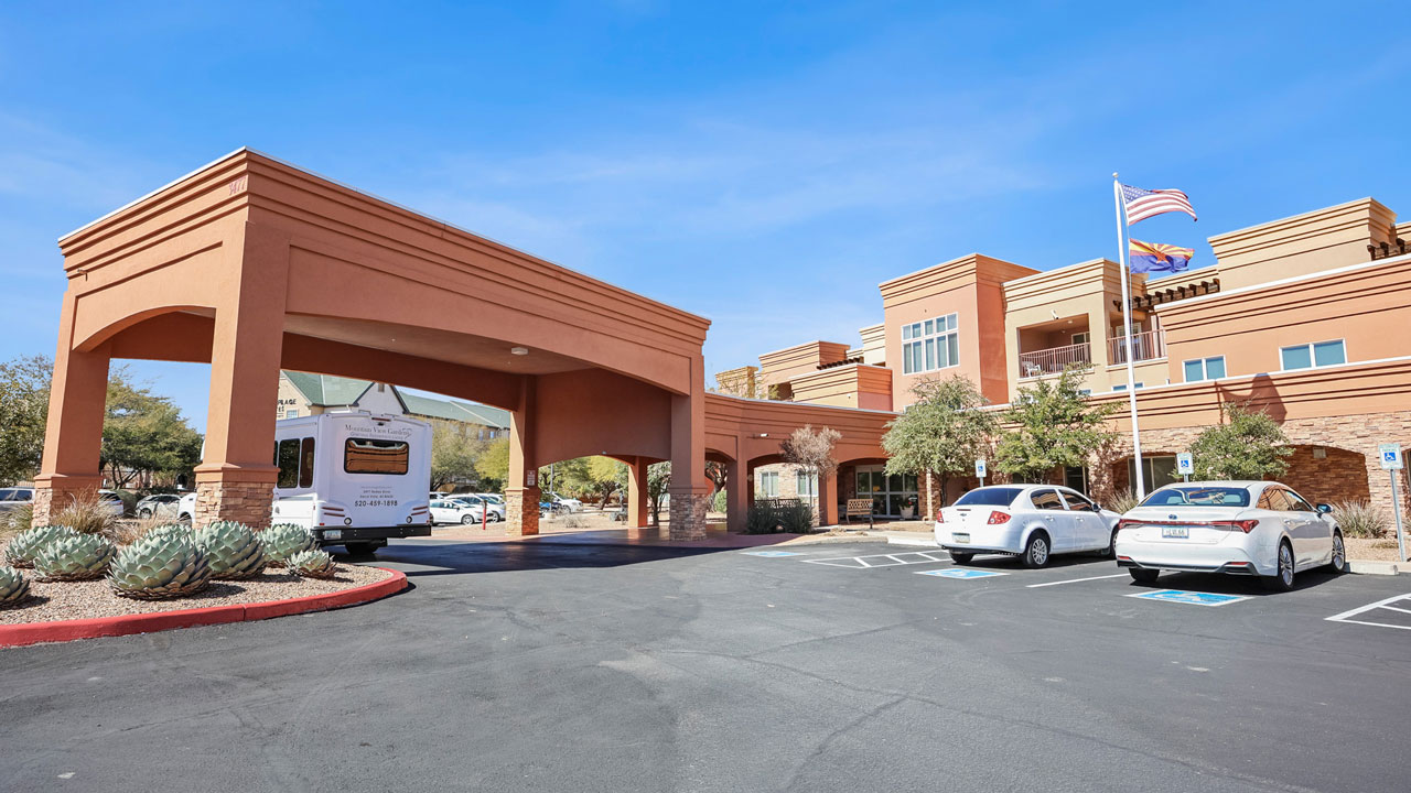 A large building with an arched drive-through entrance, several parked cars, a flagpole with American and state flags, and landscaped desert plants under a clear blue sky.