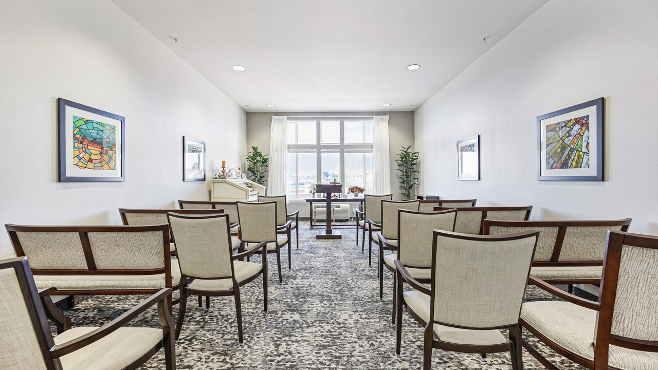 A small, bright room with rows of beige chairs facing a window. Artwork hangs on white walls, and two plants stand near the window. A piano and refreshment area are visible at the front of the room.