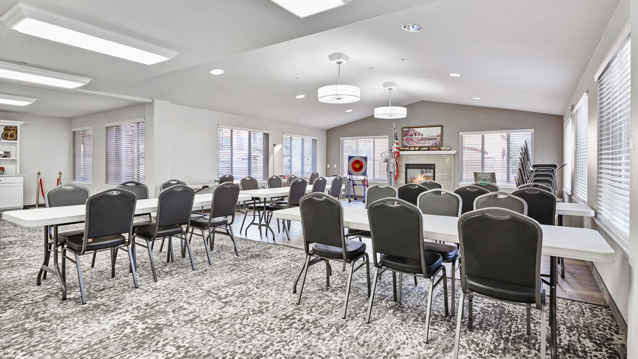 A spacious, well-lit meeting room with several rows of black chairs and white tables facing a fireplace, American flag, and wall decor. Large windows let in natural light.