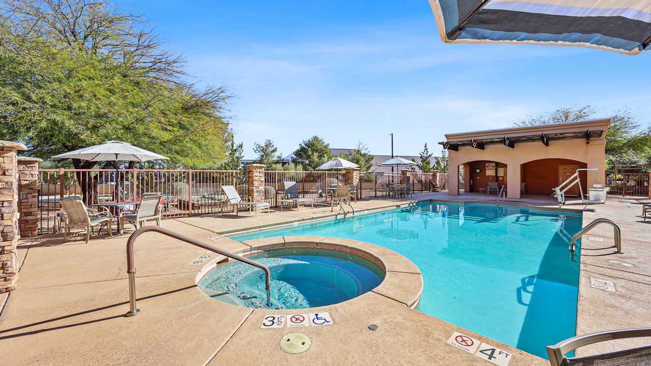 Outdoor swimming pool area with a hot tub, lounge chairs, umbrellas, and shaded seating. The pool has a step entry, accessible features, and is surrounded by a fence and trees under a clear blue sky.