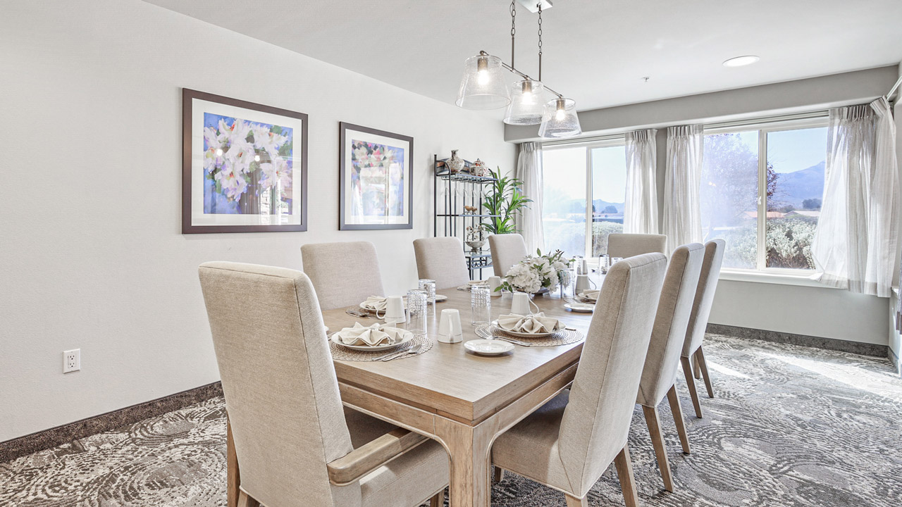 A bright dining room with a wooden table set for six, beige upholstered chairs, floral centerpiece, wall art, a metal shelf with plants, and large windows letting in natural light.
