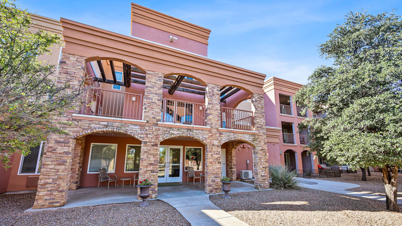 Two-story building with stone arches, terracotta walls, balconies, and large windows, surrounded by trees and landscaped gravel paths under a clear blue sky.