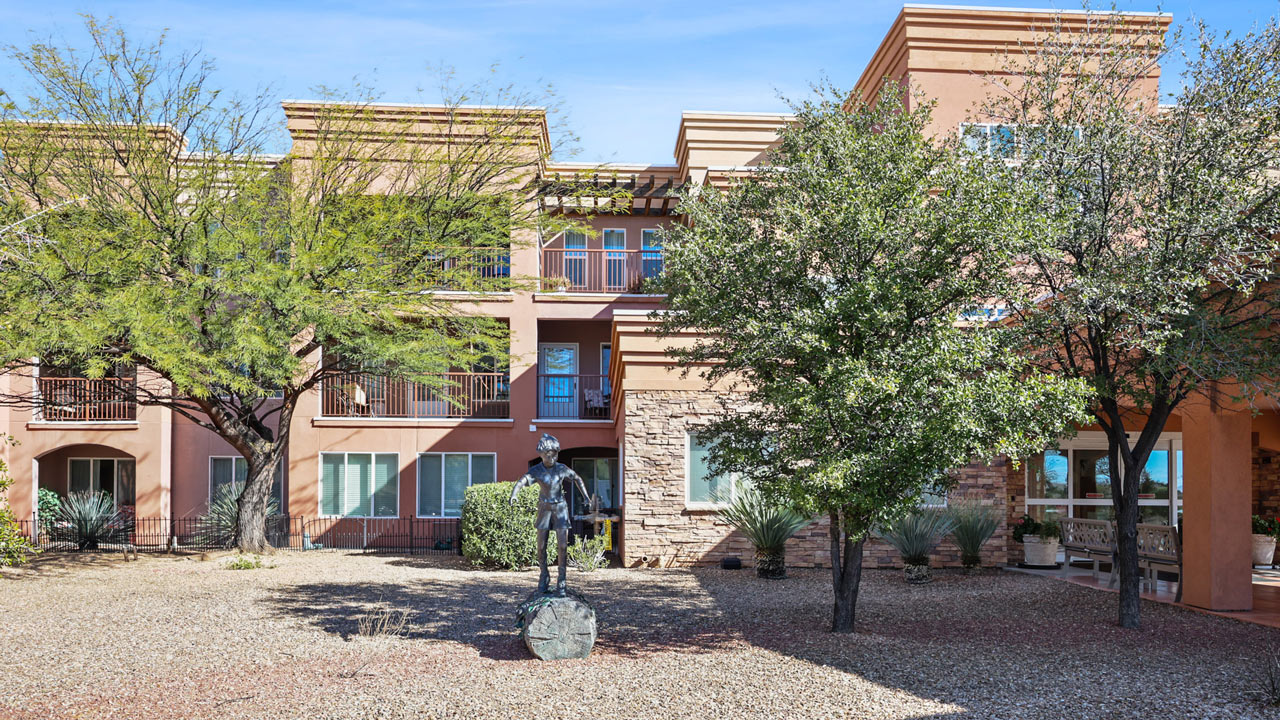 Three-story tan building with balconies, surrounded by trees and desert landscaping. A metal statue of a person riding a horse is centered in the courtyard. Clear blue sky overhead.