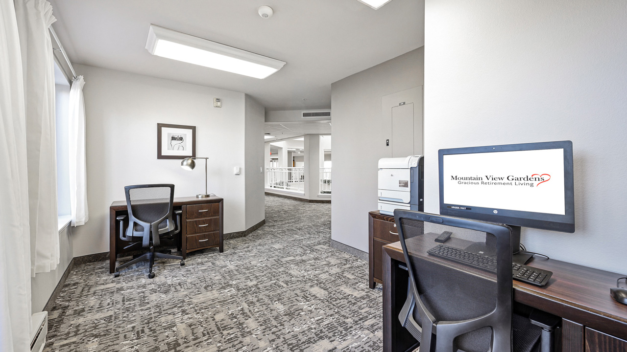 A modern office space with two desks and chairs, one with a computer displaying "Mountain View Gardens Gracious Retirement Living," gray carpet, white walls, and bright overhead lighting.