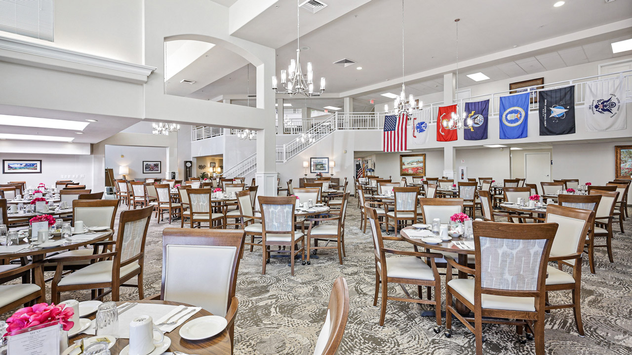 A spacious, well-lit dining hall with many wooden tables and chairs, set for a meal. Chandeliers hang from the ceiling, and military flags and the U.S. flag are displayed on a balcony railing.