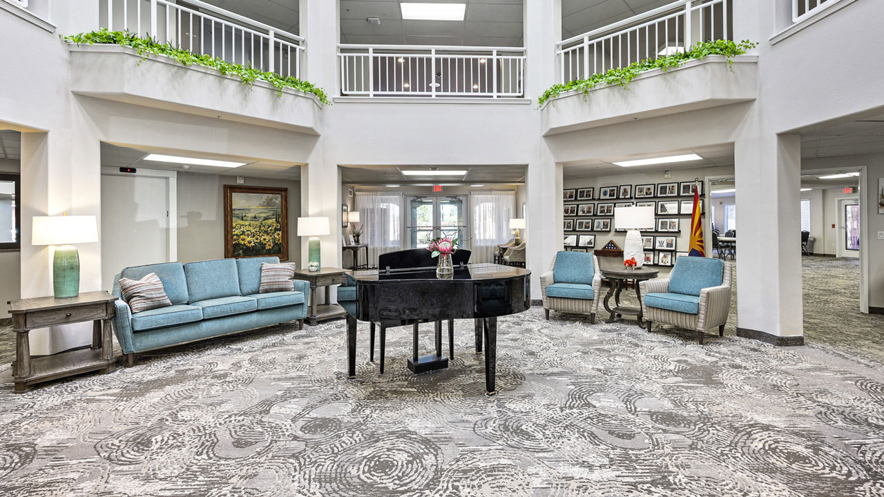 A spacious, elegant lobby with a black grand piano in the center, surrounded by comfortable blue sofas and armchairs, modern decor, plants on the upper balcony, and framed photos on the far wall.