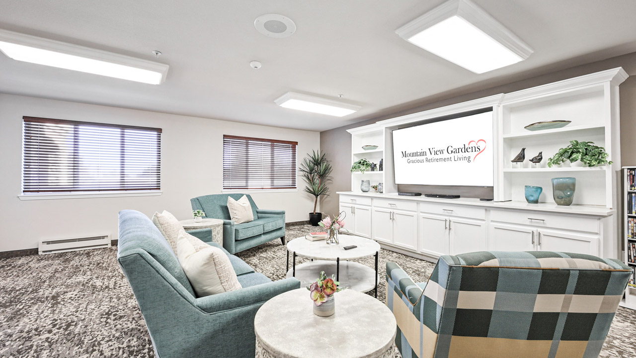 A bright, modern lounge features teal armchairs, a checkered chair, round tables with plants, and a large TV displaying "Mountain View Gardens: Gracious Retirement Living" on a white wall unit with shelves and decor.