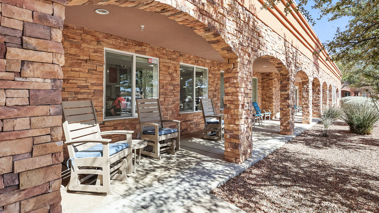 A covered stone patio with several cushioned wooden rocking chairs faces large windows, bordered by arches, with gravel landscaping and desert plants.