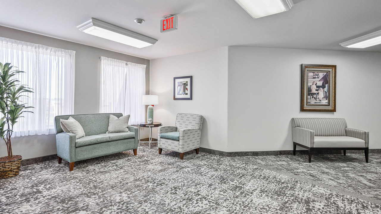 A bright, modern waiting room with two windows, light curtains, a green loveseat, two armchairs, a floor lamp, a potted plant, framed art on white walls, and gray patterned carpet.