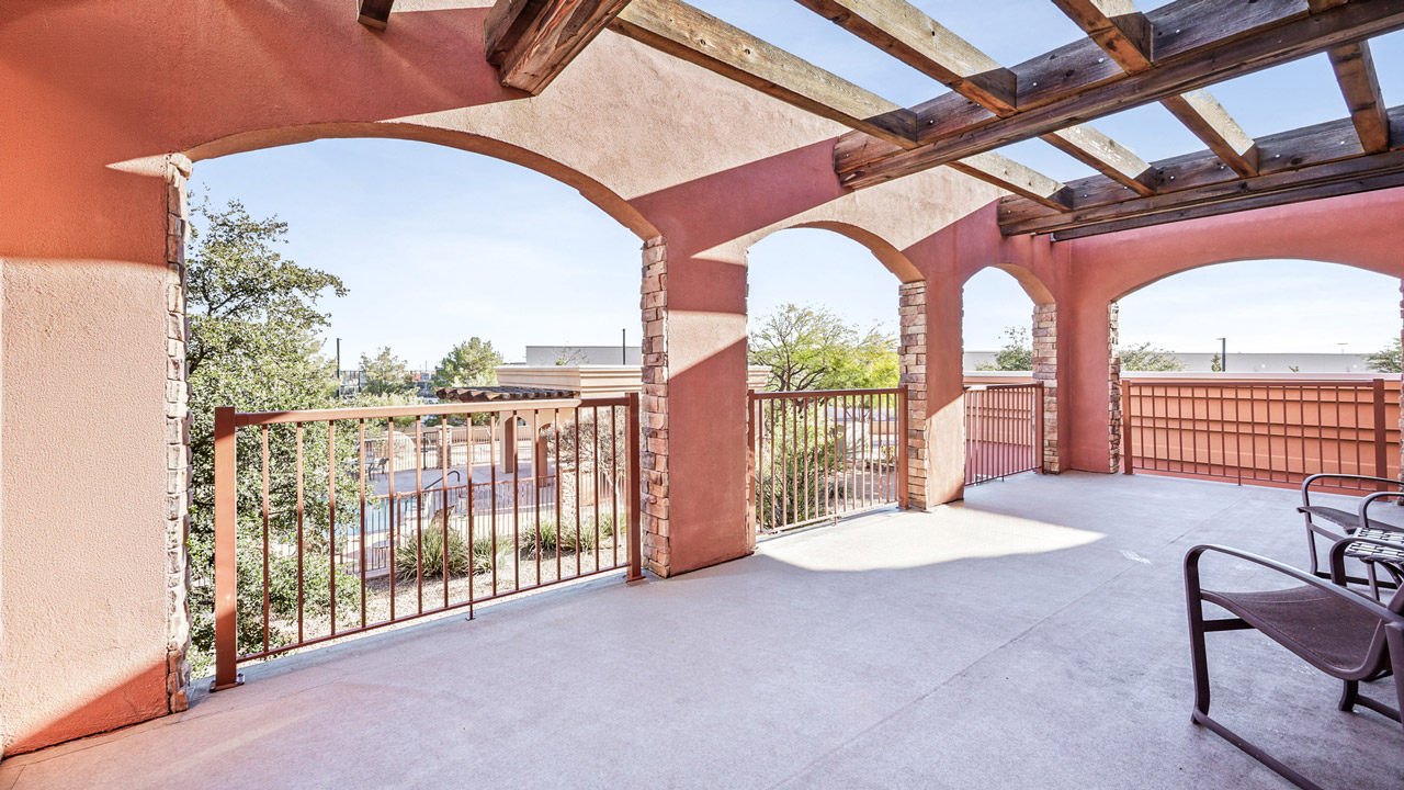 A spacious balcony with a wooden pergola, arched openings, metal railings, and outdoor chairs, overlooking trees and buildings on a sunny day.