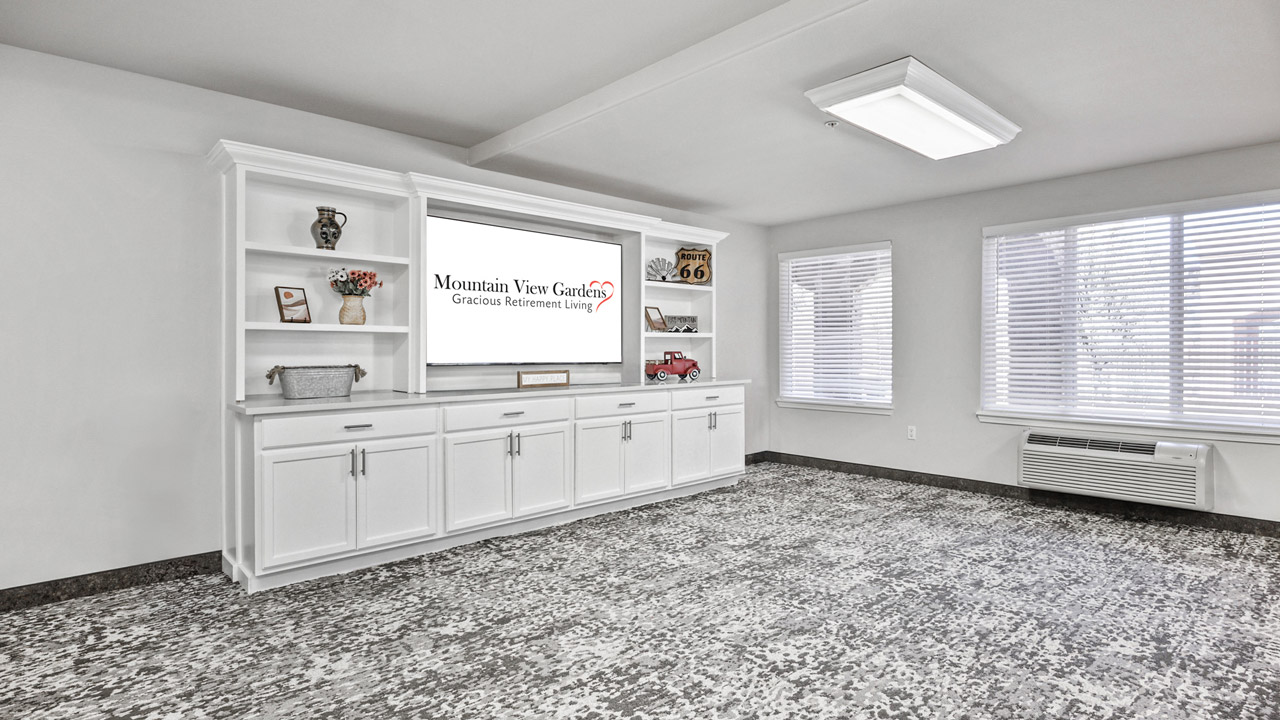 A bright, modern room with white built-in shelves and cabinets, a large TV displaying "Mountain View Gardens Gracious Retirement Living," patterned carpet, and two windows letting in natural light.