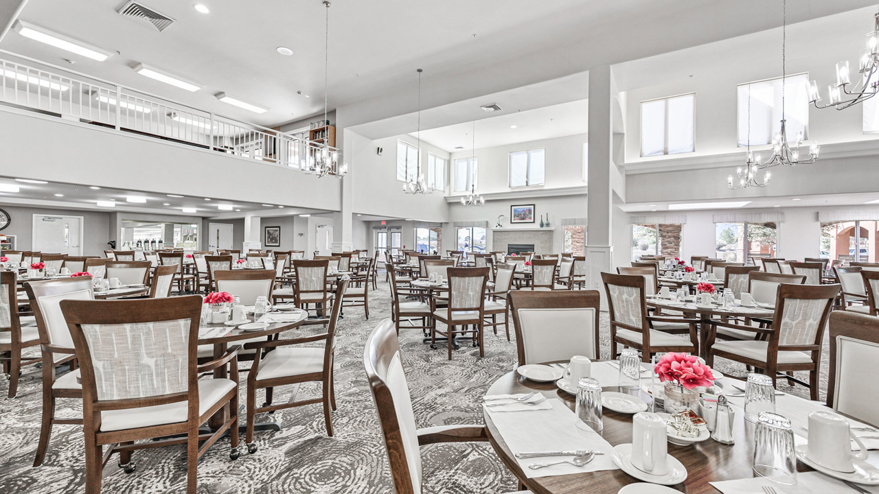 Spacious dining hall with many wooden tables and chairs, set with white tableware and bright pink flower centerpieces. Large windows and chandeliers provide ample natural and artificial light. The overall atmosphere is clean and elegant.