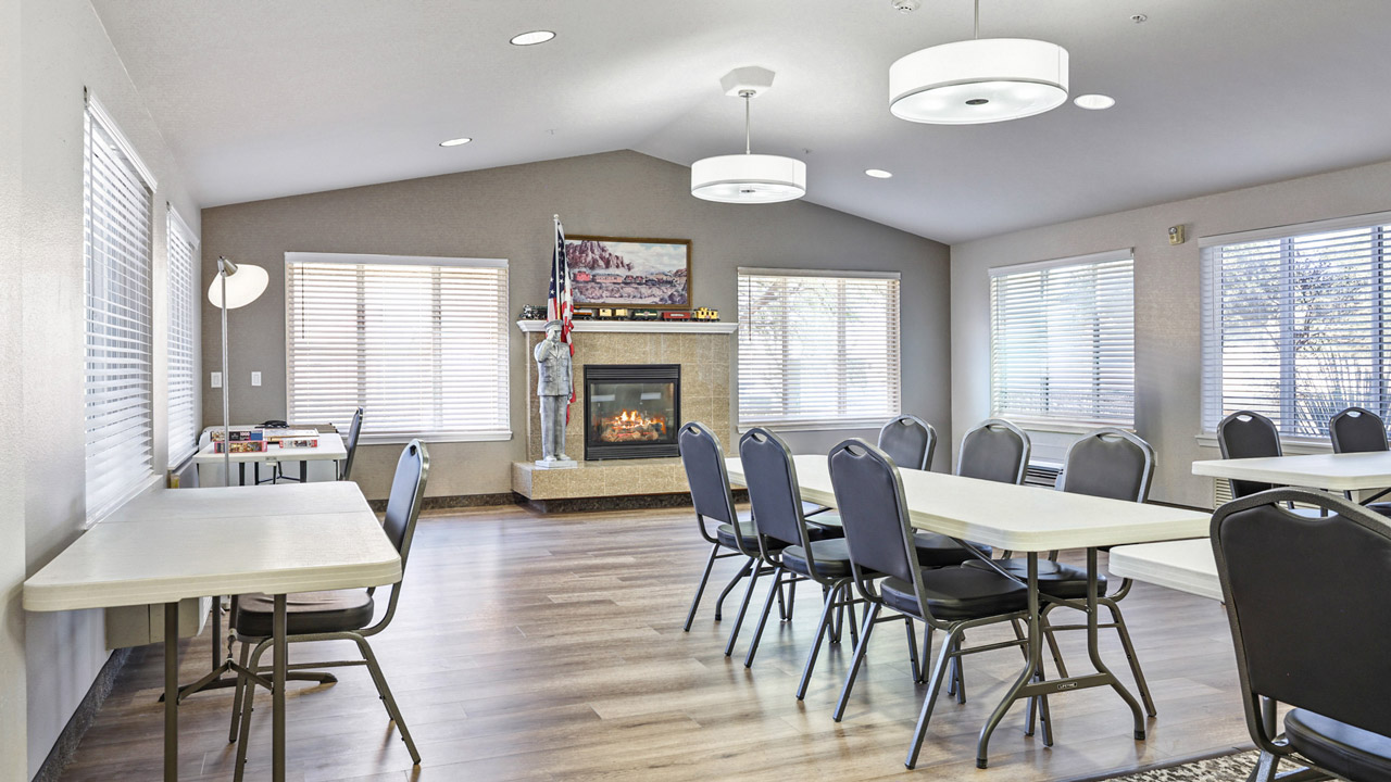 A bright room with several rectangular tables and black chairs, wood flooring, large windows, a cozy fireplace, and a TV mounted above the mantel. Light fixtures hang from the ceiling.