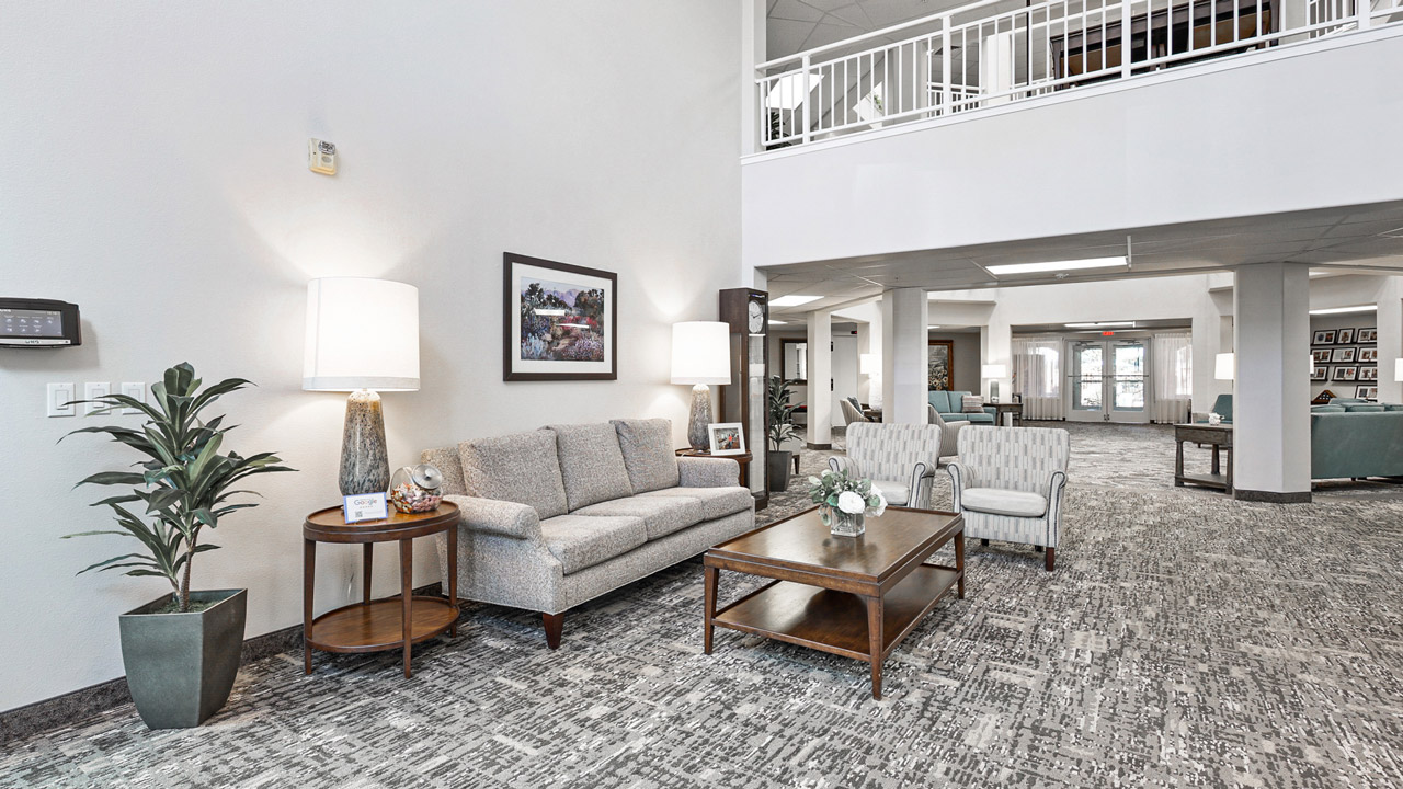 A modern, bright lobby with a gray sofa, two patterned armchairs, wooden coffee and side tables, table lamps, a potted plant, and framed art on the wall, open to a second floor with white railings.