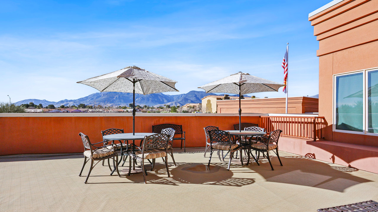 Outdoor patio with two round tables, each shaded by an umbrella, surrounded by metal chairs. In the background are mountains, clear blue sky, and an American flag on a pole.