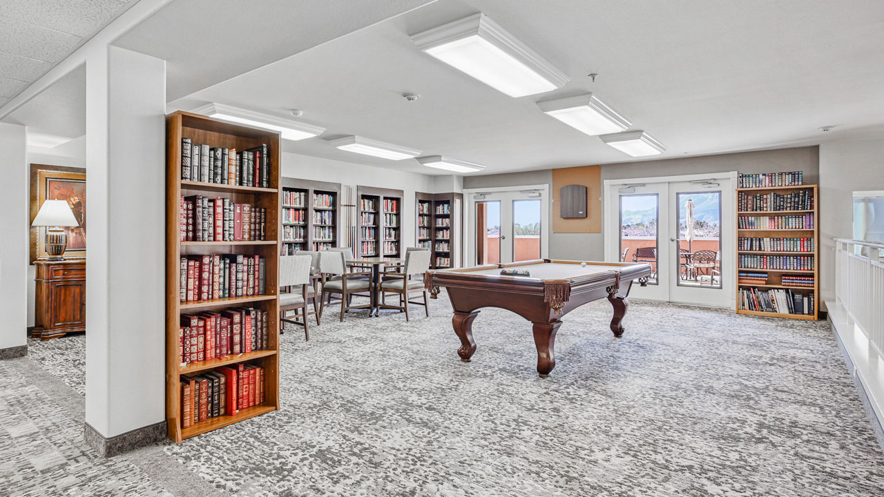 A bright library room with bookshelves lining the walls, a wooden pool table in the center, a small table with chairs, and large windows letting in natural light with a view outside.