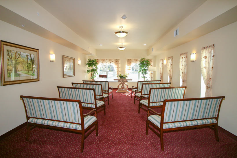 A small chapel with red carpeting, rows of striped cushioned benches, framed paintings on the walls, and large windows covered with sheer curtains. The room includes potted plants and a table with flowers at the front.