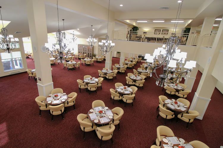 A spacious dining hall with red carpeted floors and white walls is filled with round tables covered with white cloths, set for a meal. Elegant chandeliers hang from the high ceiling, and there is a mezzanine level with a beige railing.