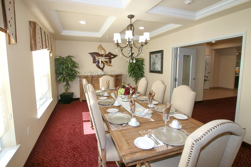 A well-lit dining room with beige walls and a red carpet features a long wooden table set for eight. The table is adorned with plates, cups, and napkins. The room includes cushioned chairs, a chandelier, decorative plants, and wall art.