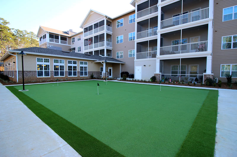 A residential building with multiple balconies overlooks a well-maintained green putting area with small flags. The building has beige walls, white trim, and several windows. The area is bordered by paved walkways and landscaped with small plants.