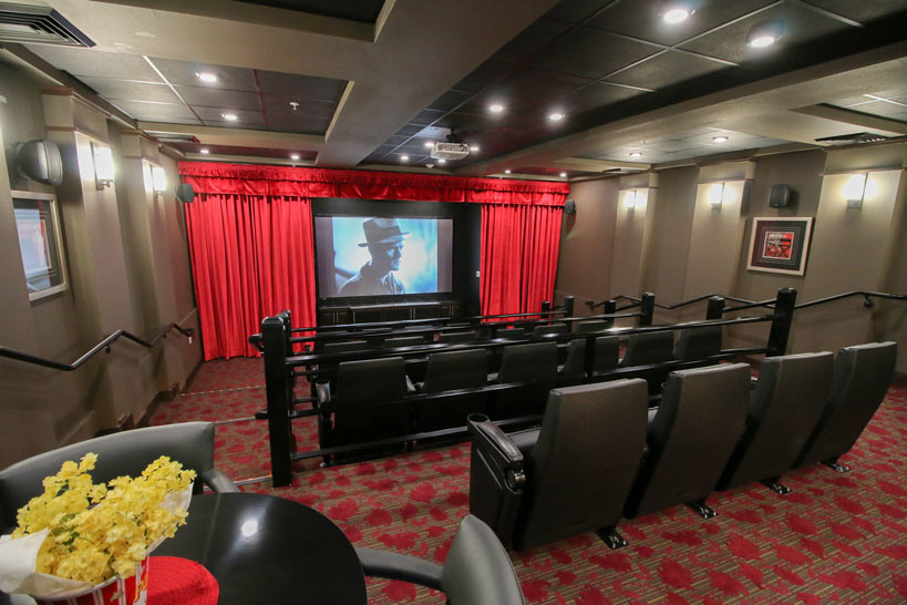 A small, modern movie theater with red carpet flooring and gray upholstered seats arranged in rows. A large screen displays an image of a person wearing a hat. Red curtains frame the screen. A table with a popcorn bucket is in the foreground.