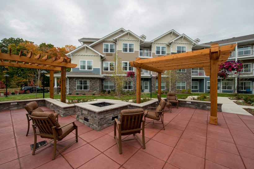 A well-maintained patio with wooden pergolas, several chairs, and a central fire pit in front of a multi-story residential building. The sky is overcast, and hanging flower baskets add touches of color to the scene.