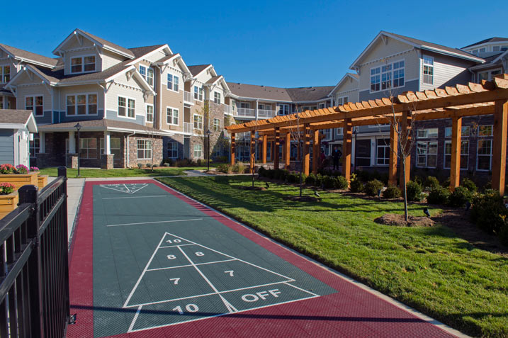 A multi-story residential building with beige and brown siding features an adjacent outdoor recreation area. The courtyard includes a shuffleboard court, a pergola with wooden beams providing shaded seating, and landscaped green spaces. Clear skies are above.