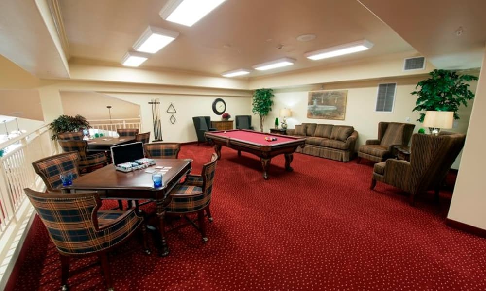 A cozy game room with red carpet features a pool table at the center, surrounded by a set of brown sofas and armchairs. On the left, a wooden table with four checkered chairs is arranged for cards or board games. The room is warmly lit with ceiling lights.