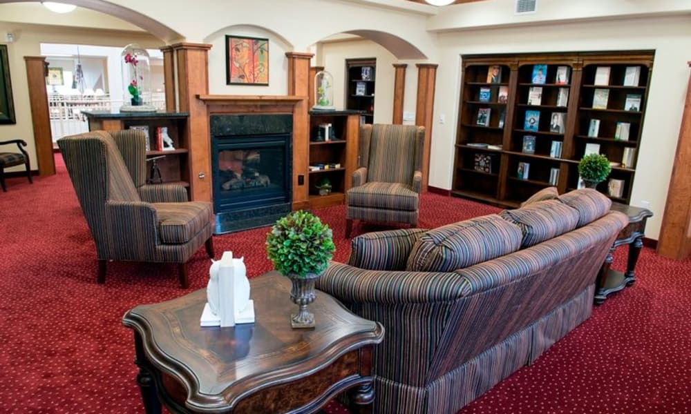 A cozy library room with a fireplace, surrounded by wooden bookshelves filled with books and decor items. There are comfortable striped armchairs and a matching sofa arranged around a wooden coffee table with decorative plants. The room has red carpet flooring.