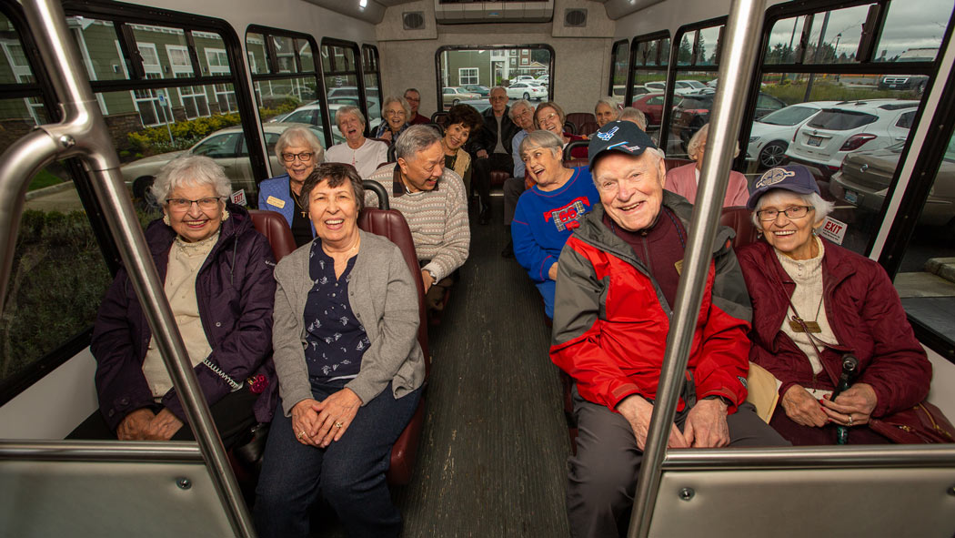 A group of elderly individuals is seated inside a bus, smiling and appearing cheerful. The bus is parked, and the large windows show a view of parked cars and a building exterior. The passengers are dressed in casual clothing and seem to be enjoying their time together.