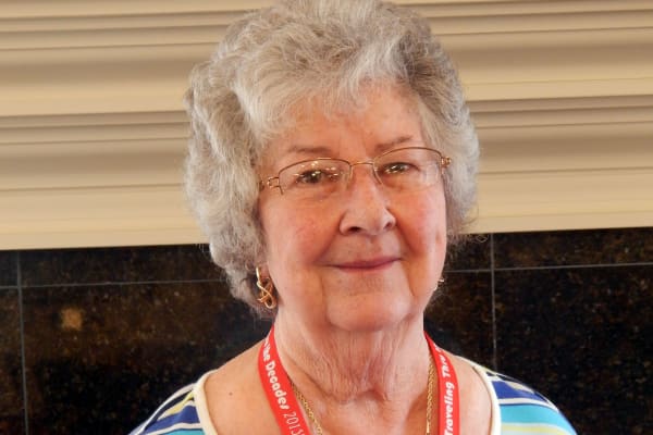 An elderly woman with short, curly gray hair and glasses wears a red lanyard around her neck. She is dressed in a white and blue striped top and smiles pleasantly while standing indoors against a dark and light-colored wall background.