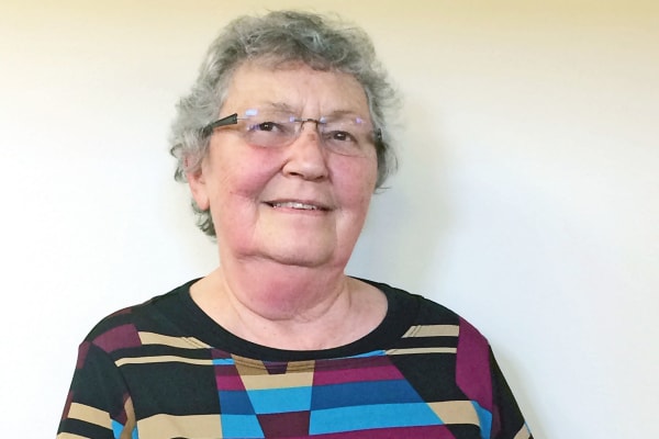 An elderly woman with short, curly gray hair and glasses smiles softly at the camera. She is wearing a colorful geometric-patterned top with shades of red, yellow, blue, and black. The background is a plain light-colored wall.