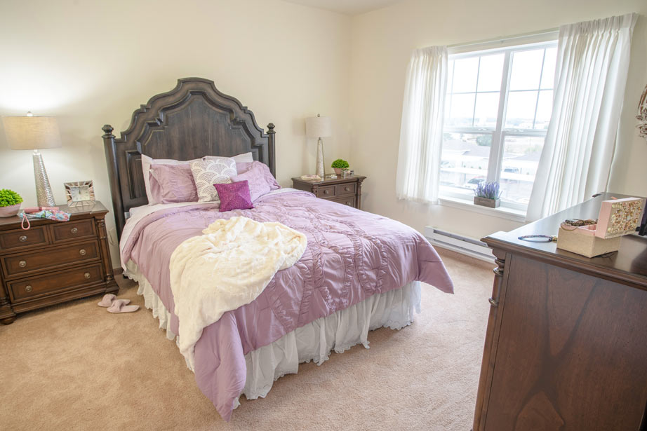 A cozy bedroom featuring a dark wood bed with a high, ornate headboard, dressed in a lavender comforter and white bed skirt. The room includes matching nightstands with lamps, a dresser with decor items, and a window with white curtains letting in natural light.