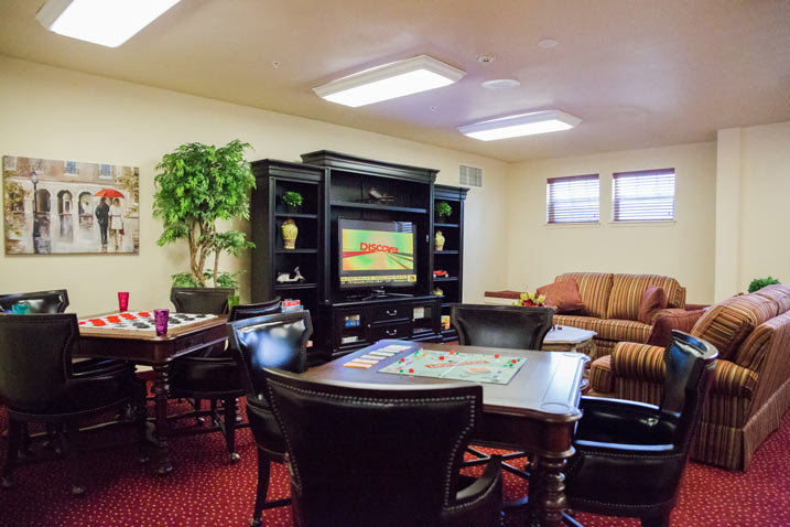 A cozy lounge room features board game tables with games set up, several couches and armchairs, a large entertainment center with a TV displaying 