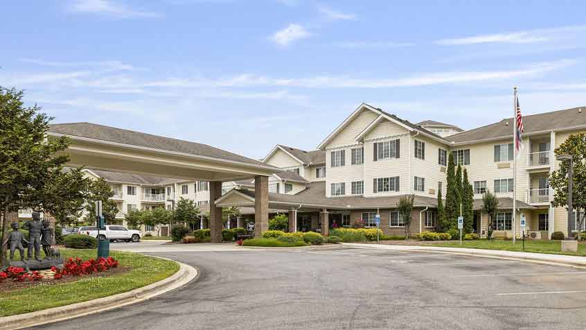 A large, two-story cream-colored building with multiple windows, an entrance canopy, landscaped greenery, a circular driveway, and a flagpole, likely a residential or retirement community, under a blue sky.