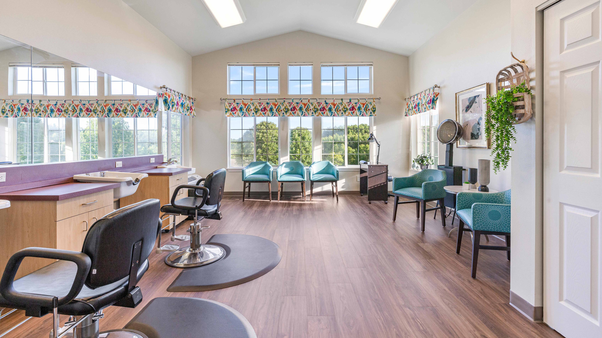 A bright, modern salon with styling chairs, mirrors, a row of teal waiting chairs by large windows, wood floors, and colorful valances. Greenery and wall art add decorative touches to the inviting space.