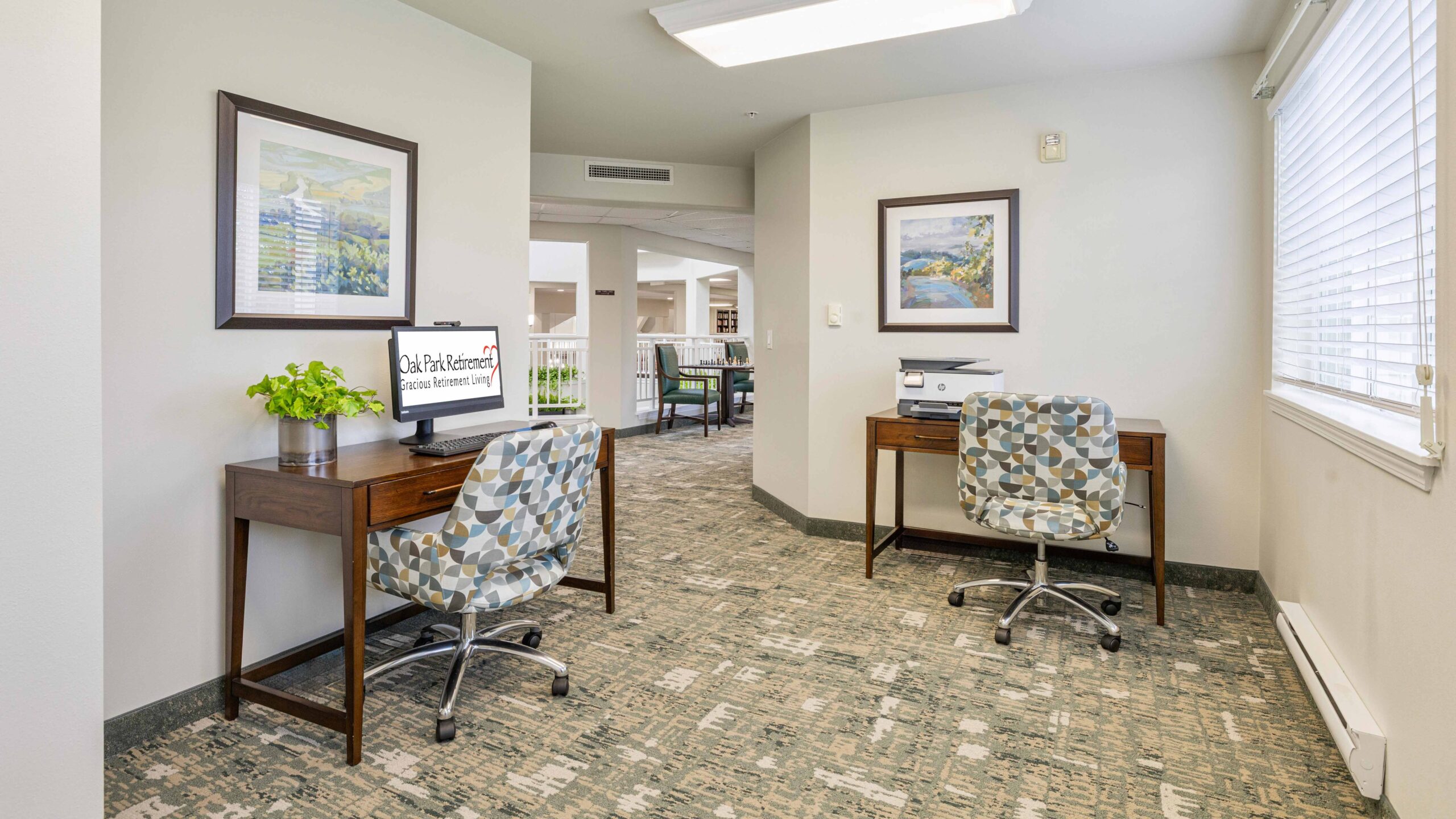 A small office area with two wooden desks, each with a patterned chair. One desk has a computer and plant, the other has a printer. Bright space with art on the walls and a window letting in natural light.