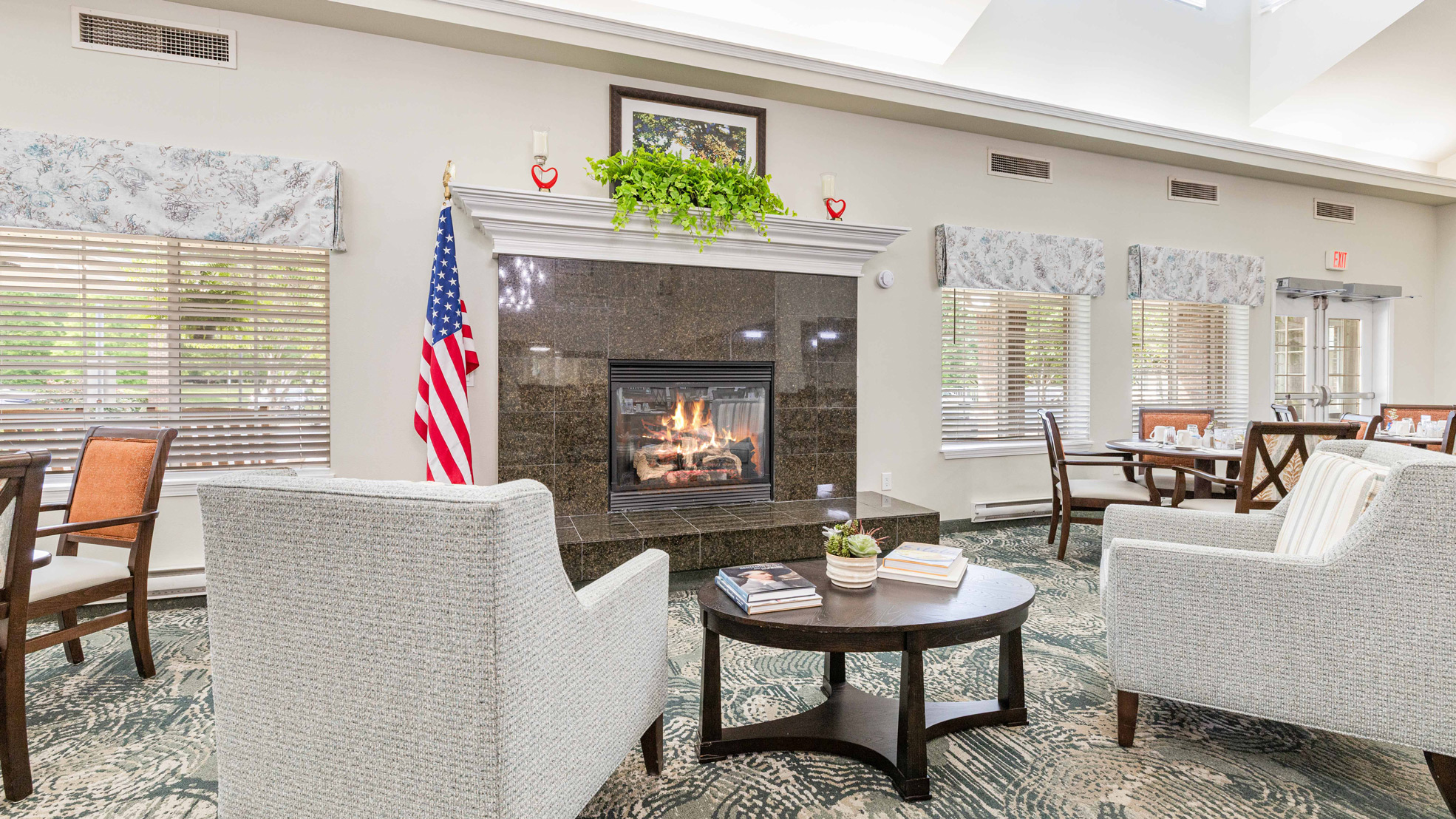 A bright, spacious lounge with two armchairs facing a lit fireplace, an American flag beside it, and large windows with floral valances. A round table with books sits between the chairs on a patterned carpet.