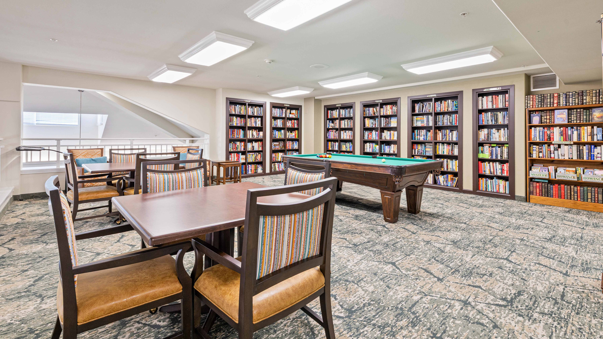 A bright room with bookshelves lining the back wall, a pool table in the center, and two wooden tables with chairs. The room has patterned carpet and modern ceiling lights.