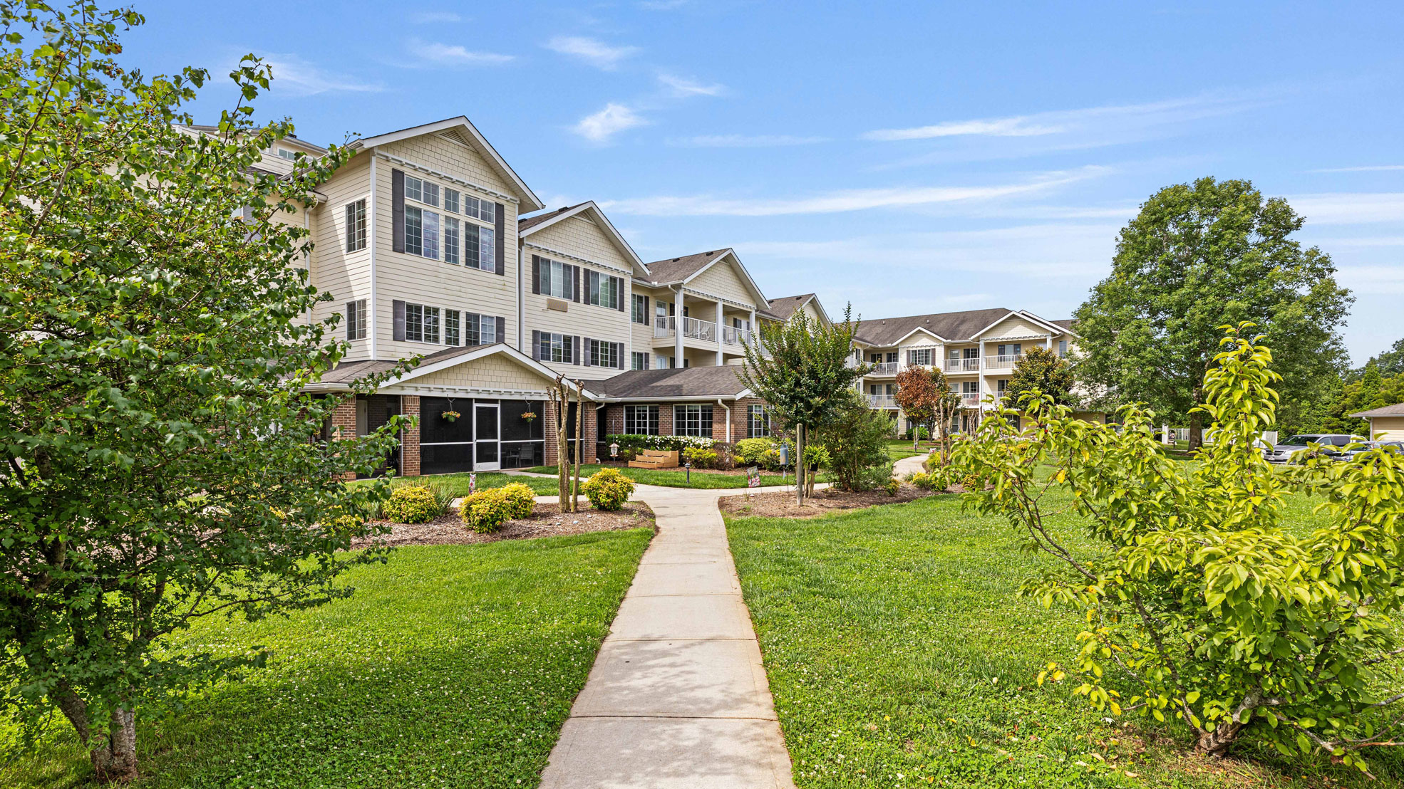 A sidewalk leads to a large, three-story apartment building with white siding and many windows, surrounded by green grass, trees, and landscaped shrubs under a blue sky with light clouds.