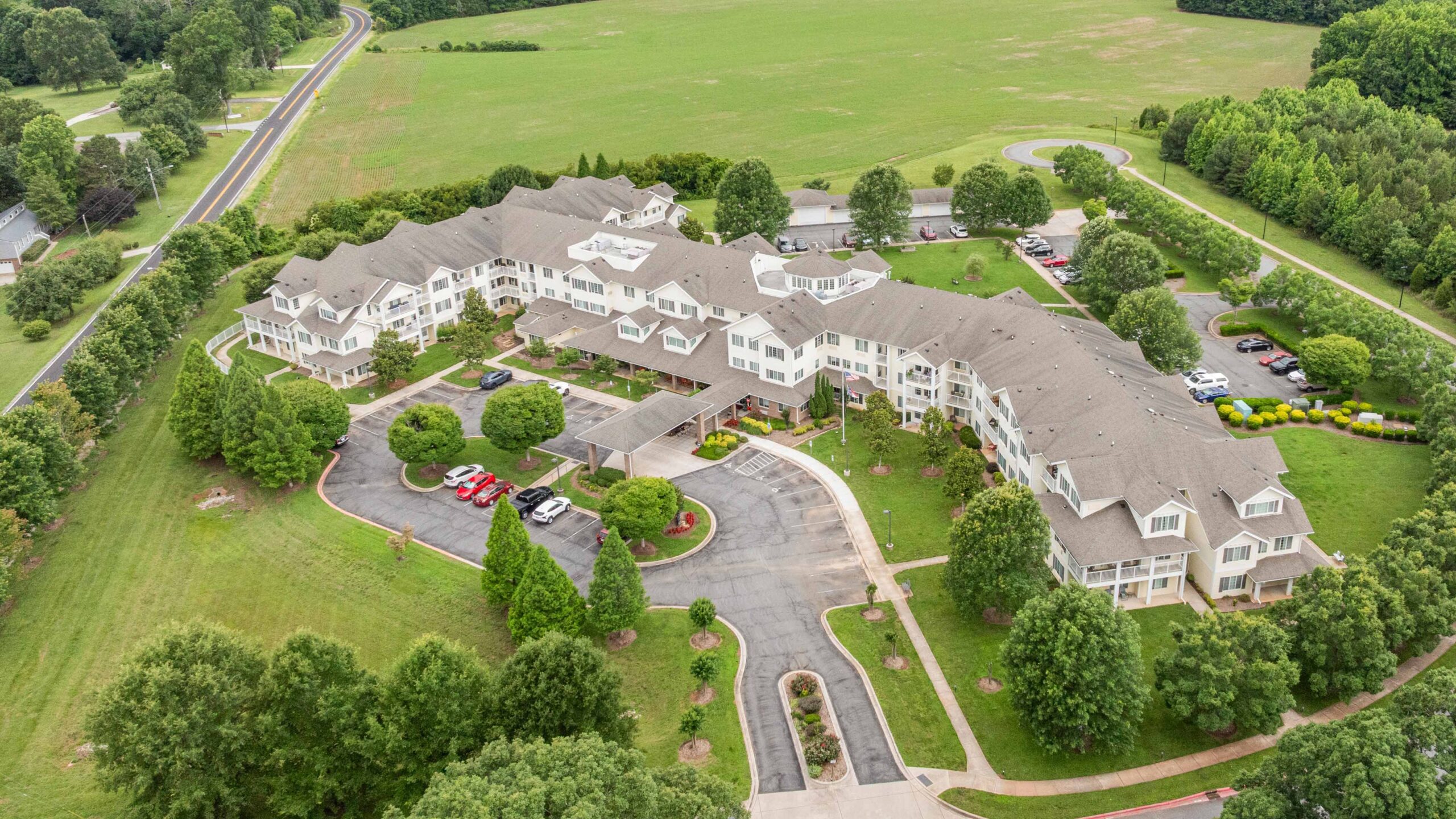Aerial view of a large, multi-story residential building with beige roofs, surrounded by green lawns, trees, parking lots, and a curved driveway, located near open fields and a winding road.