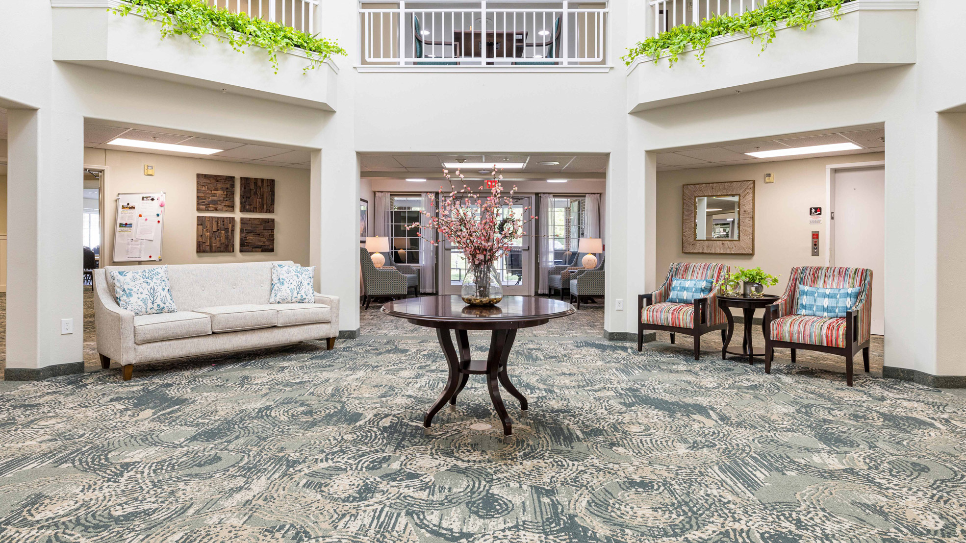 A bright, welcoming lobby with a round table holding a floral arrangement, a cream sofa with blue pillows on the left, two striped armchairs on the right, and green plants on a balcony above.