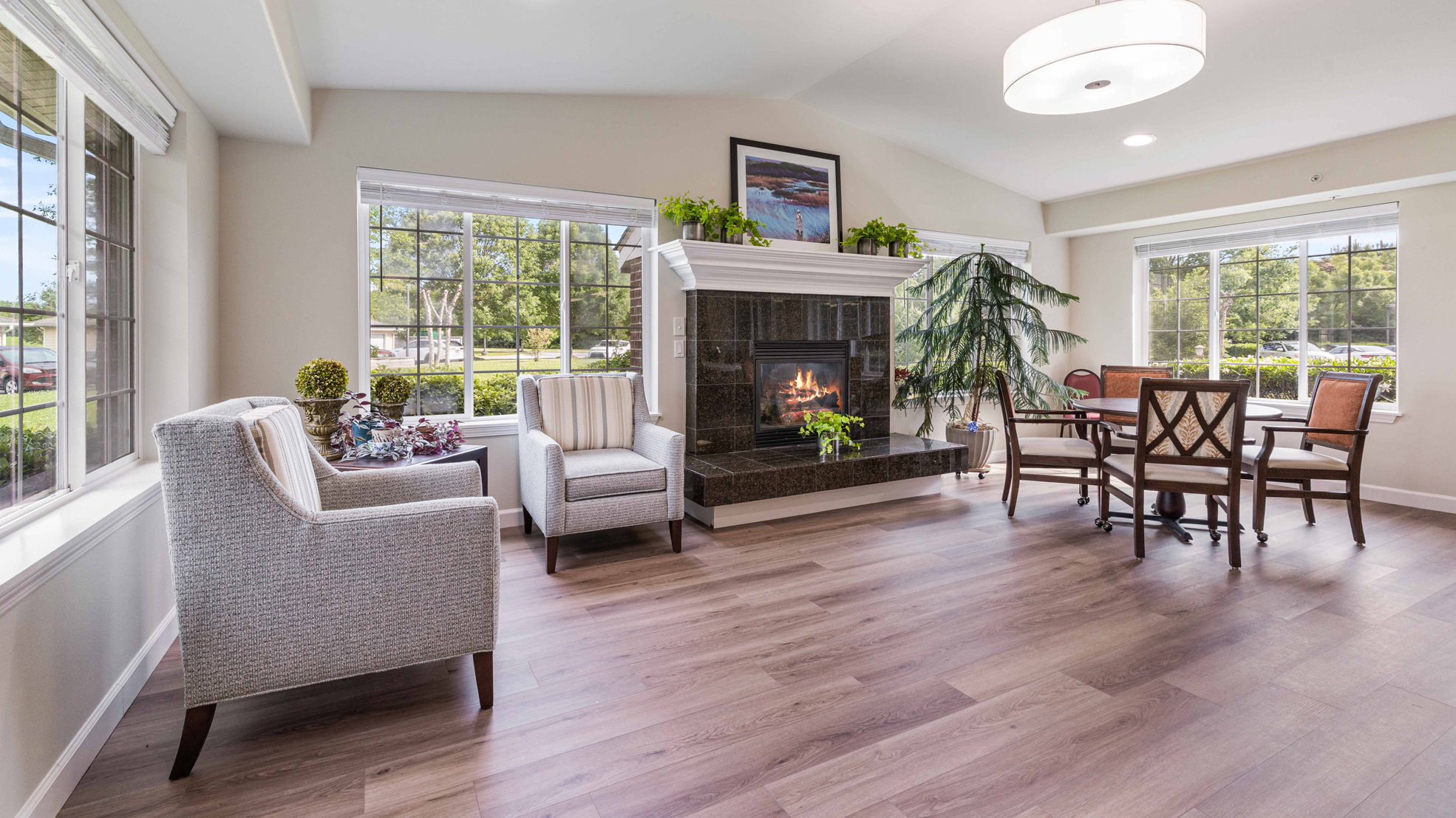 Bright living room with large windows, a fireplace, armchairs, a round dining table with chairs, houseplants, and wood flooring, all under a modern ceiling light fixture.