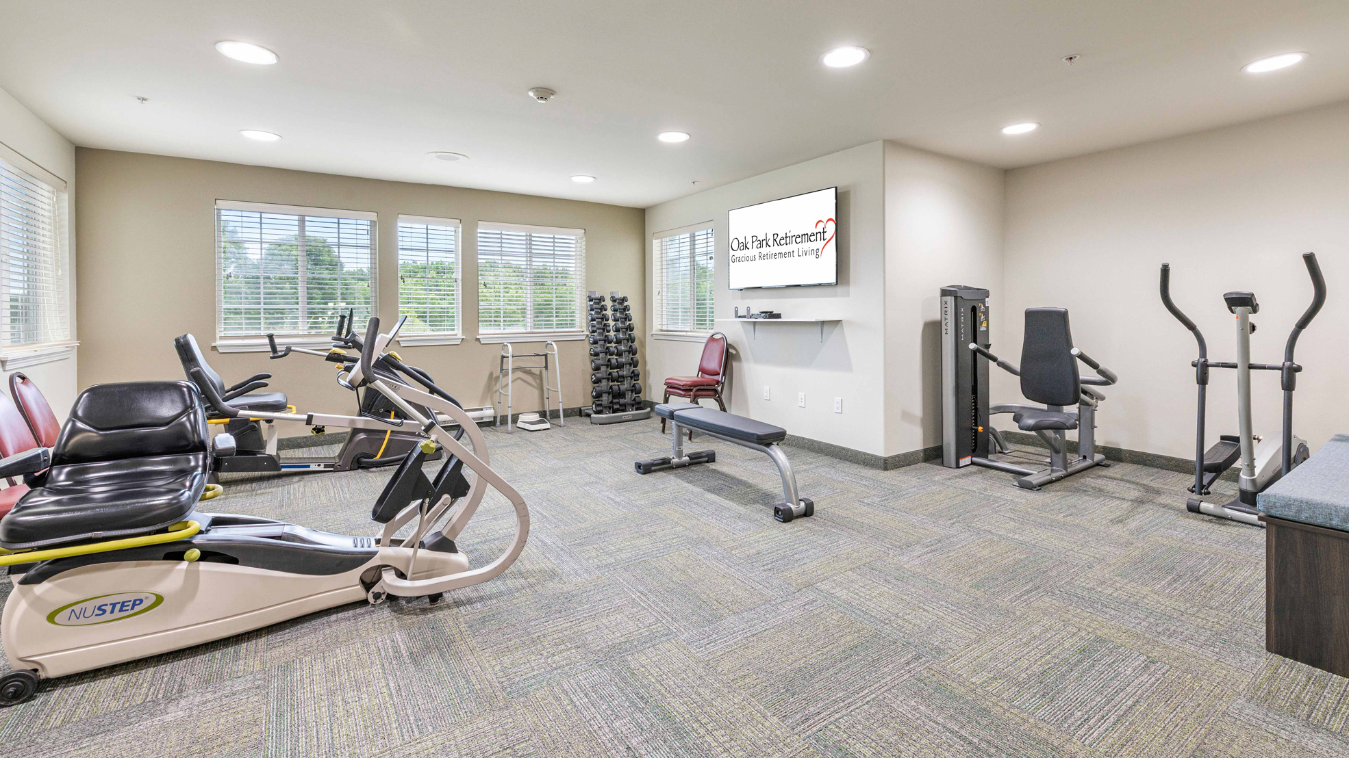A well-lit fitness room with various exercise machines, a bench, free weights, and a large wall-mounted TV displaying "Oak Park Commons." There are windows letting in natural light and carpeted flooring throughout.