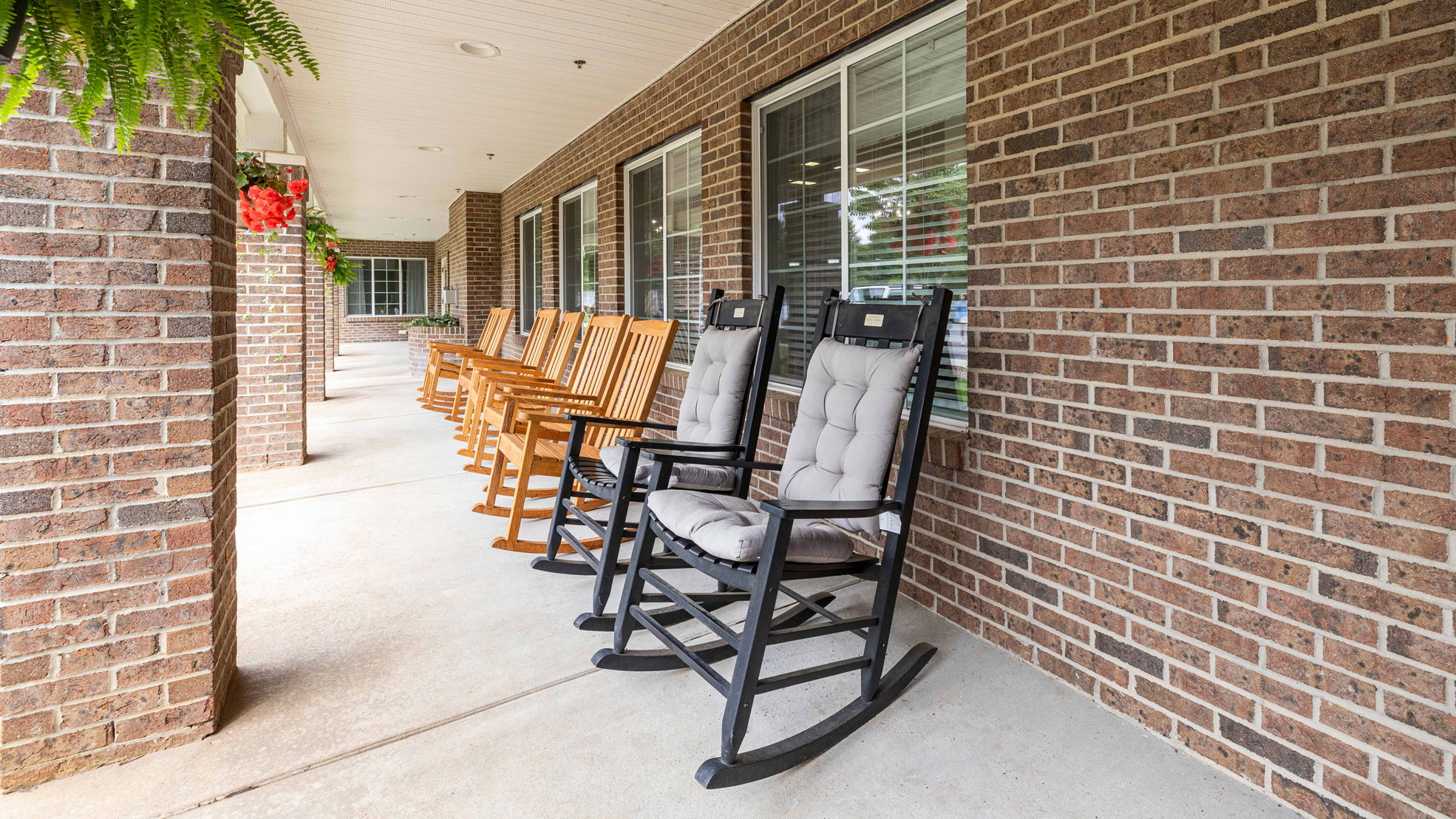 A row of wooden rocking chairs, including two with gray cushions, sits on a covered porch with brick walls and large windows. Hanging plants add greenery above the seating area.