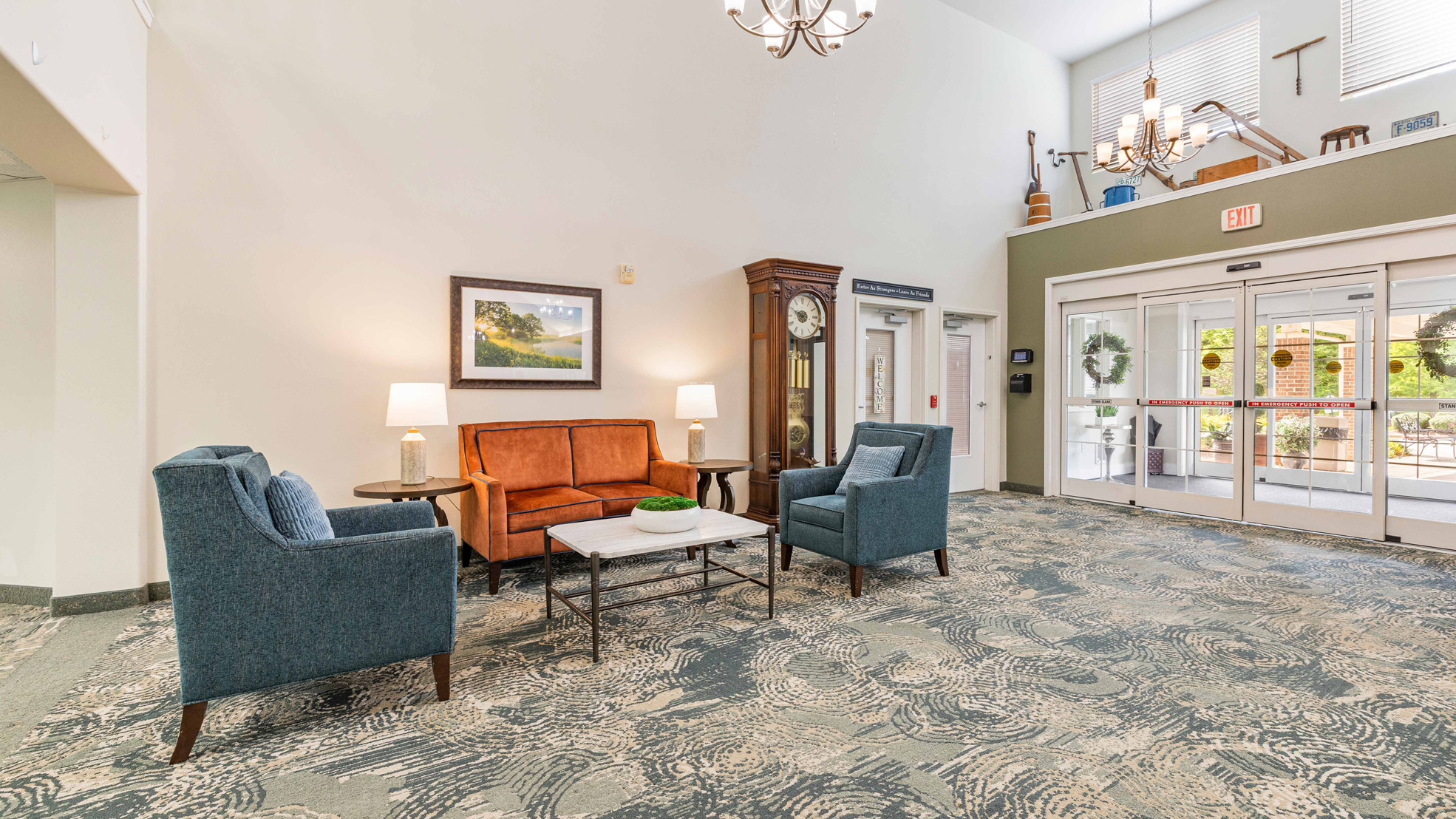 A spacious, well-lit lobby with an orange sofa, two blue armchairs around a coffee table, a grandfather clock, lamps, a large rug, and glass doors letting in natural light. Decor items are displayed on a high shelf.