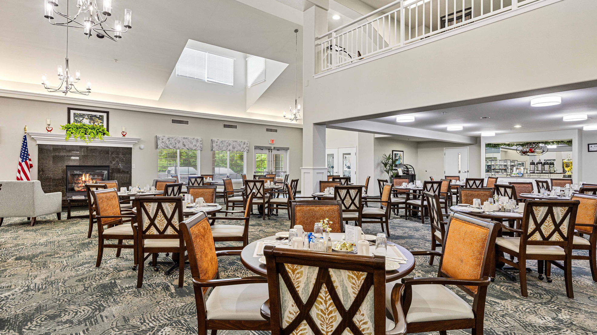 A spacious, well-lit dining room with round tables and cushioned chairs, set for meals. Chandeliers hang from the high ceiling, and a fireplace, large windows, and plants add warmth to the elegant space.