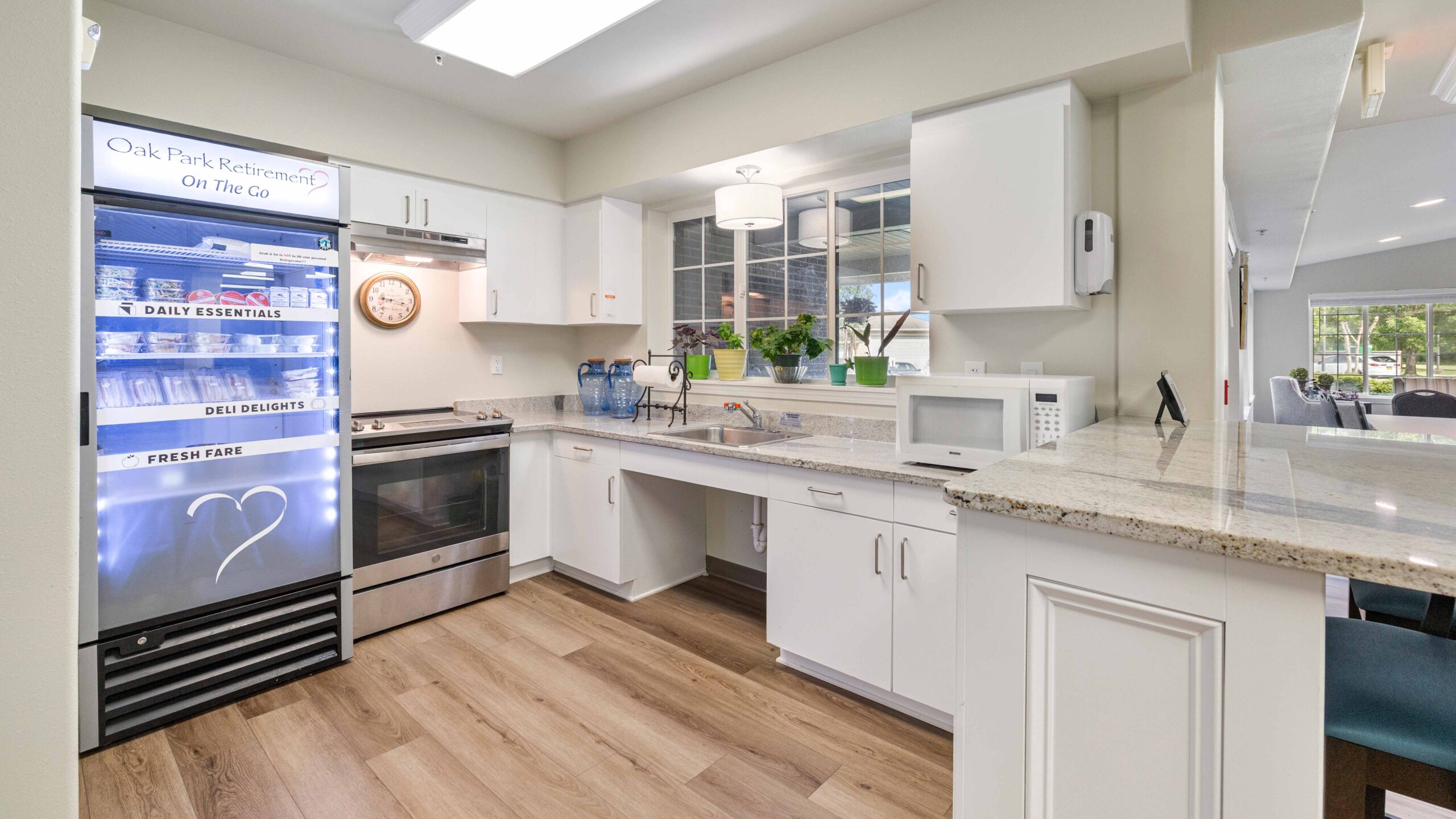 Modern kitchen with white cabinets, stainless steel appliances, a large refrigerator labeled “Oak Park Retirement On The Go,” countertop plants, microwave, and a window letting in natural light.