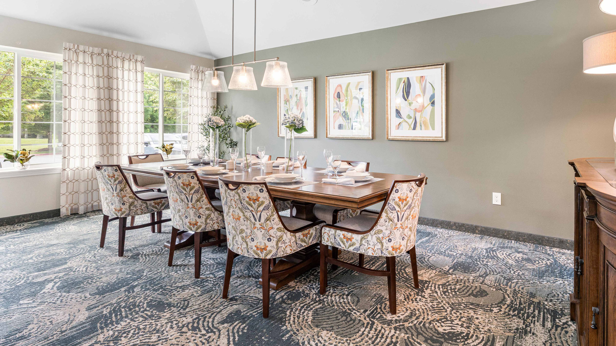 A dining room with a wooden table set for eight, patterned chairs, abstract art on a gray wall, large windows with white curtains, and blue patterned carpeting. Natural light fills the space.