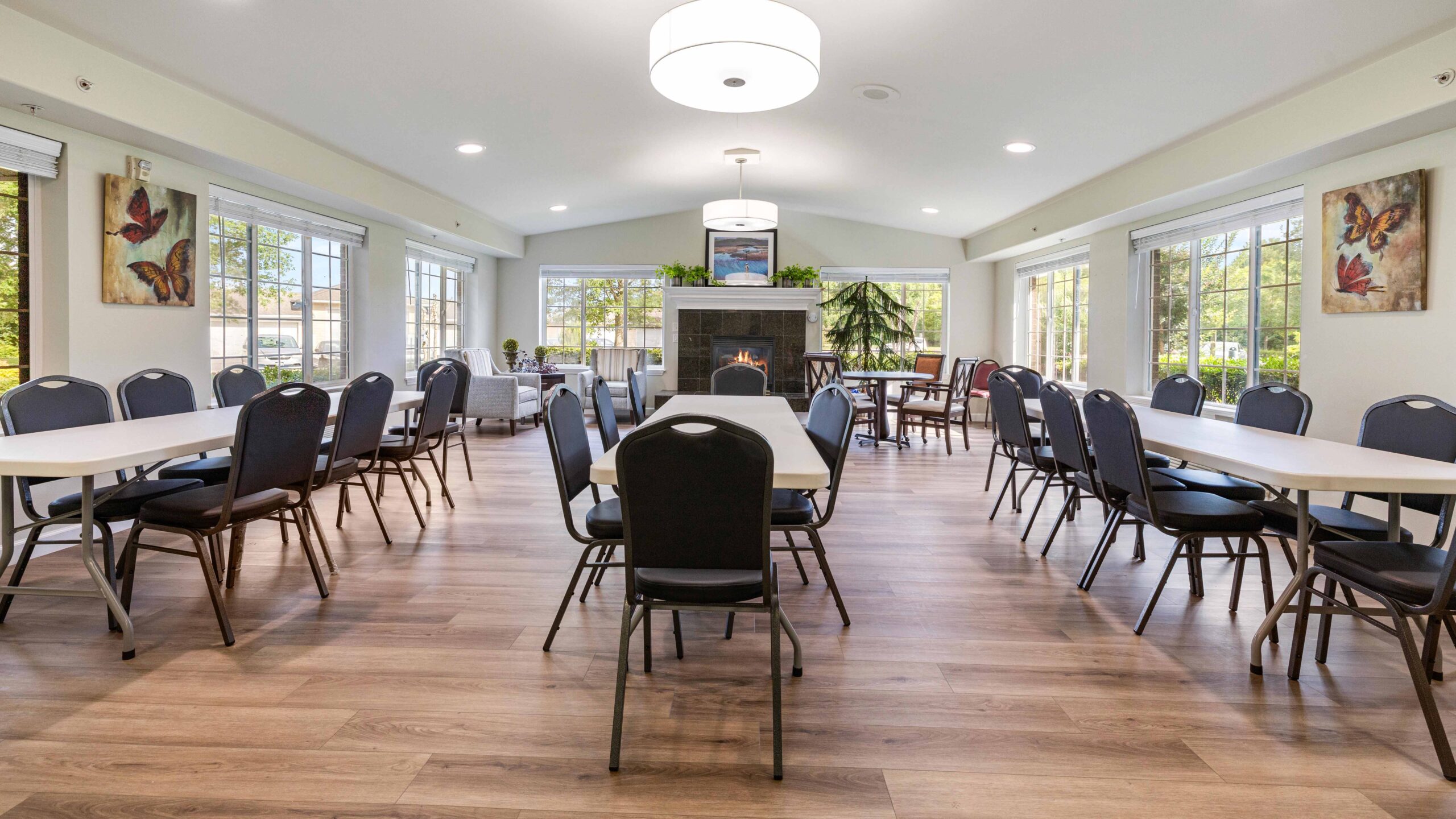 A bright, spacious dining room with several tables and chairs, wooden flooring, large windows letting in natural light, and a fireplace with plants and decor above it at the far end. Butterfly artwork hangs on the walls.